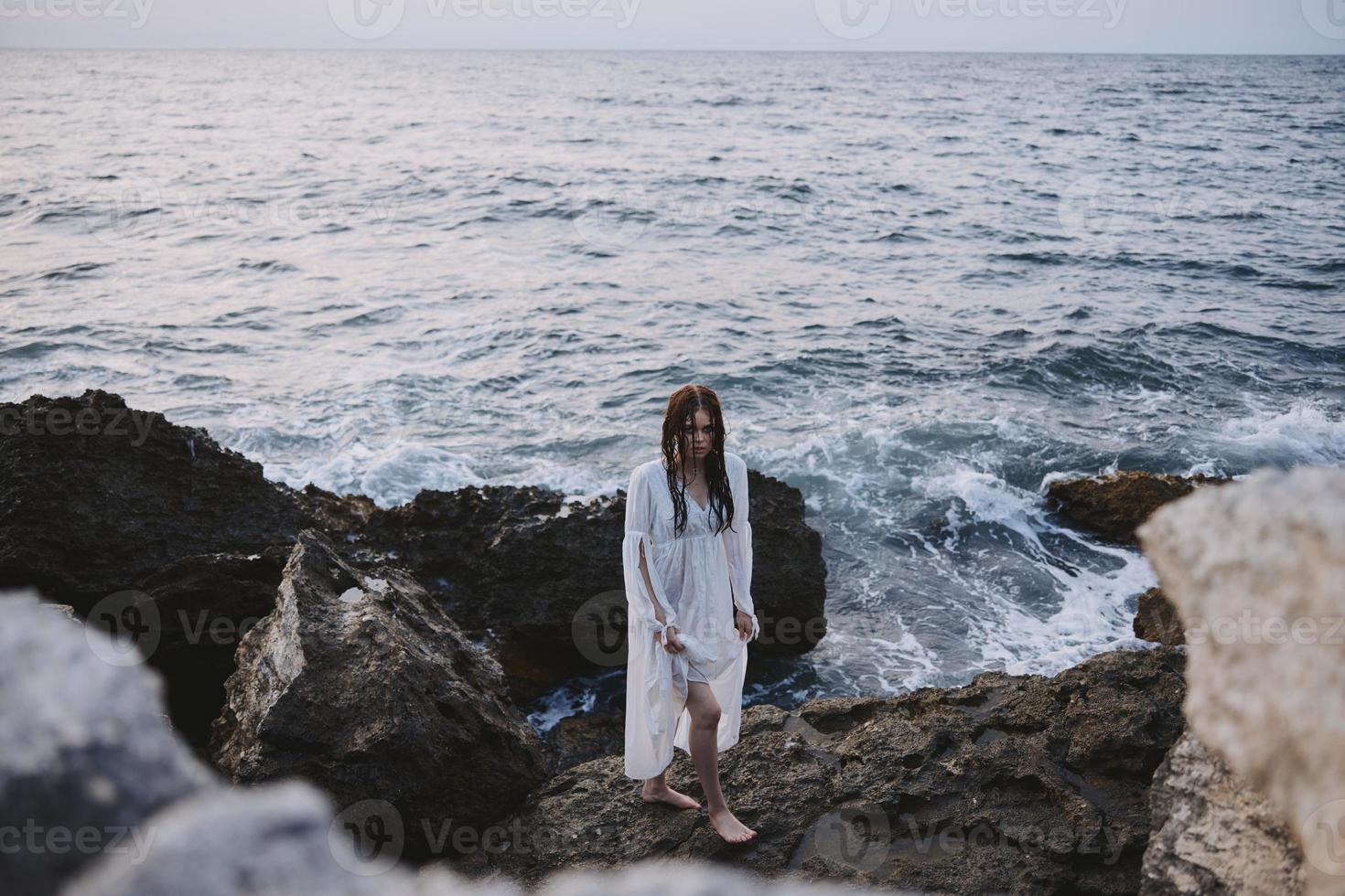 un' donna nel un' bianca vestito sta su roccioso pietre di il oceano foto