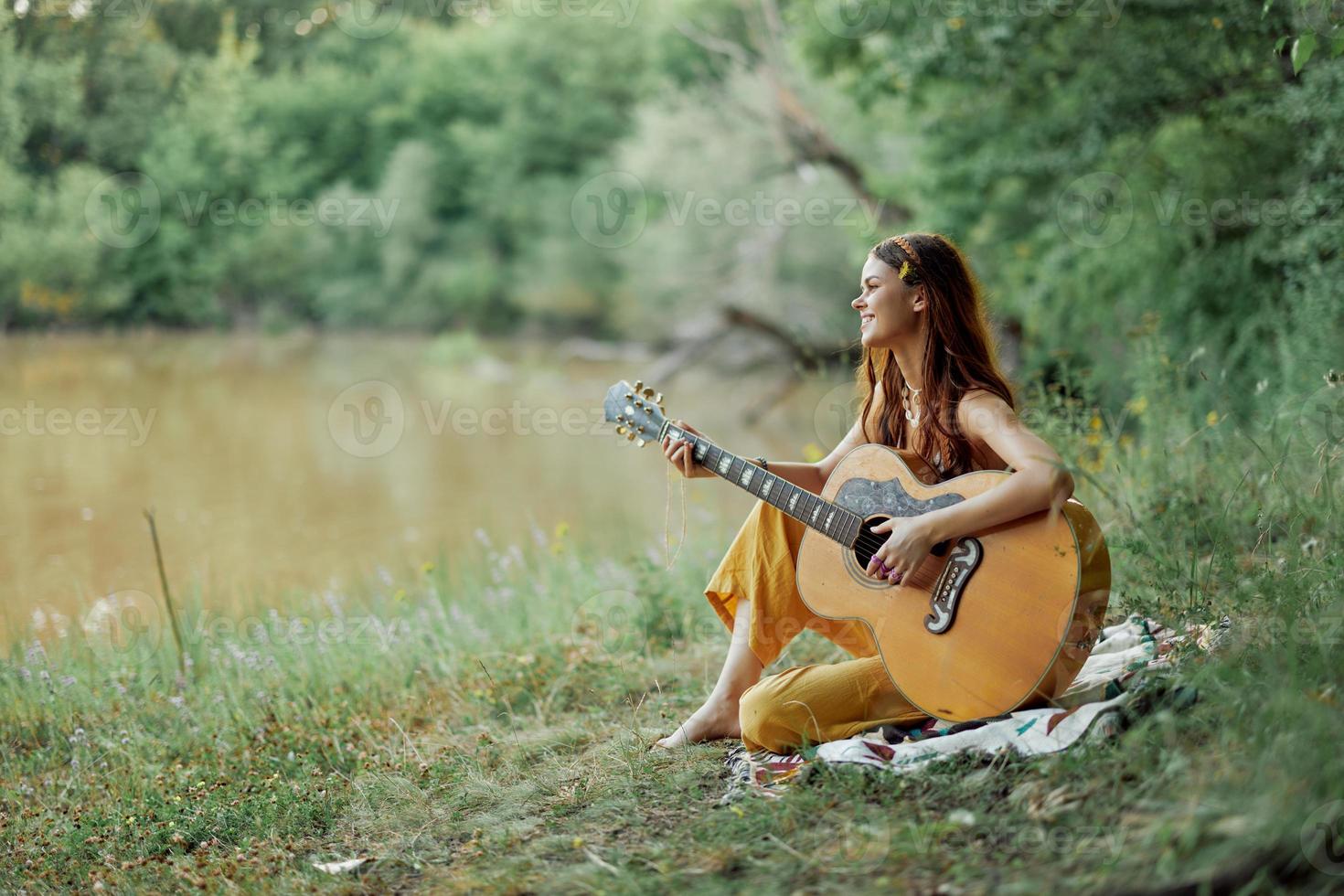 hippie donna giocando chitarra sorridente e cantando canzoni nel natura seduta su un' plaid di il lago nel il sera nel il raggi di il ambientazione sole. un' stile di vita nel armonia con il corpo e natura foto