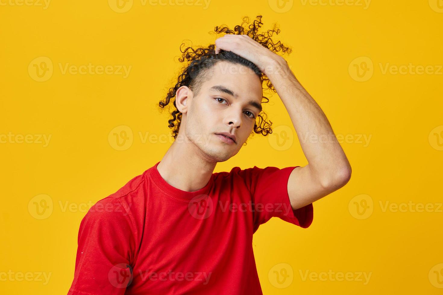 gioioso divertente caucasico giovane uomo nel rosso maglietta tocchi capelli riccioli in posa isolato su al di sopra di giallo studio sfondo. il migliore offrire con gratuito posto per pubblicità. emozioni per ogni giorno concetto foto