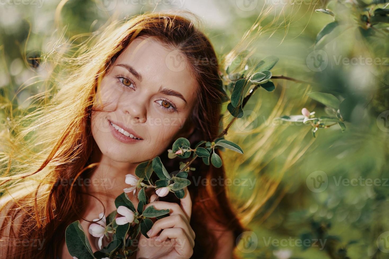 bellissimo donna Sorridi sembra a il telecamera avvicinamento con lungo rosso capelli vicino un' albero nel il estate sole nel natura nel il parco sorridente senza allergie nel un' blu vestire, il concetto di Salute e bellezza foto