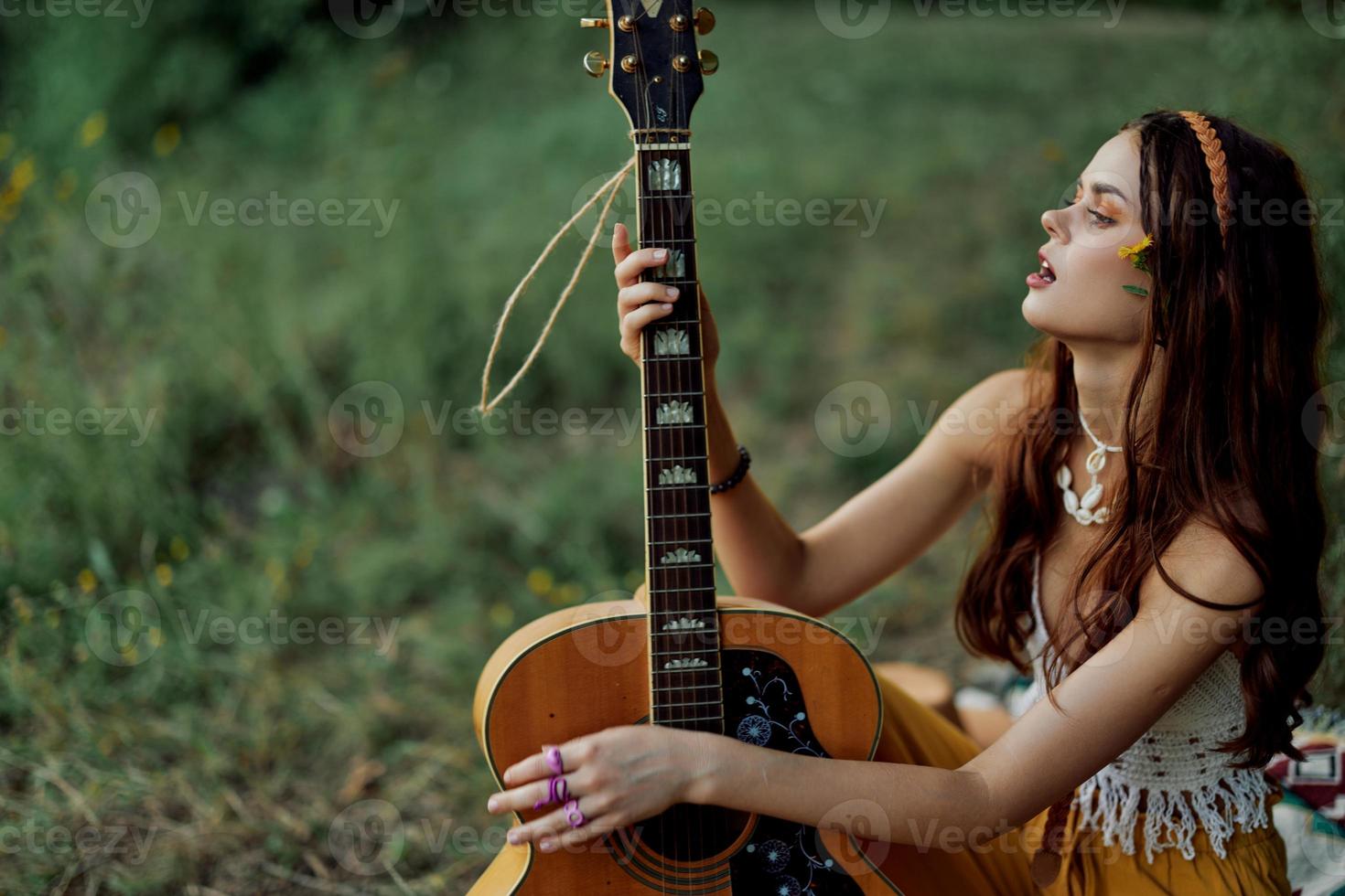 hippie eco amichevole donna con un' chitarra rilassante nel natura seduta su un' plaid di il lago nel il sera nel il raggi di il ambientazione sole. un' stile di vita nel armonia con il corpo e natura foto