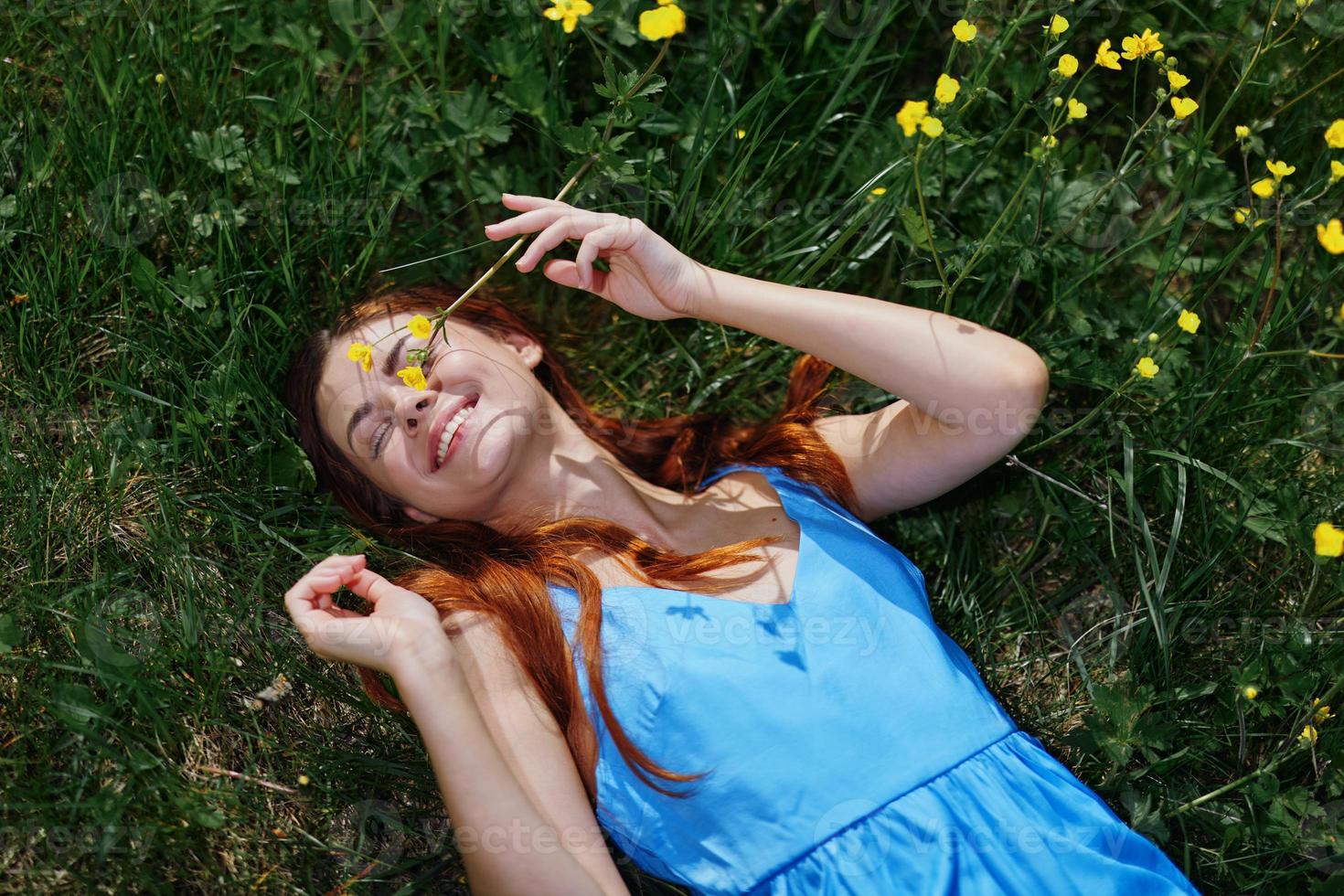 donna Sorridi con denti dire bugie su il verde erba nel un' blu vestito nel il primavera luce del sole con giallo fiori, felicità, rosso lungo capelli foto