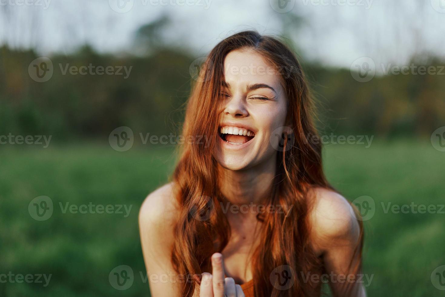 un' giovane donna sorrisi e sembra in il telecamera con sua lungo, rosso, ondulato, brillante capelli nel un' parco con verde erba nel il estate tramonto. il concetto di salutare vita, bellezza e capelli cura. foto