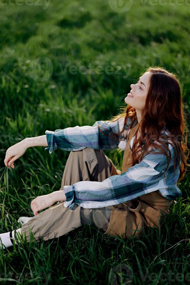 un' giovane, bellissimo donna salotti su il verde erba nel il parco indossare sneaker pantaloni e un' plaid camicia e sembra su in il ambientazione sole foto