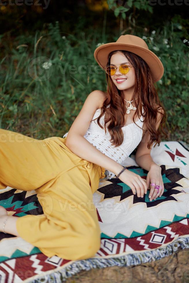 hippie donna sorridente nel eco capi di abbigliamento giallo pantaloni, bianca maglia superiore, cappello e giallo bicchieri seduta su plaid nel parco Guardando tramonto, stile di vita campeggio viaggio foto