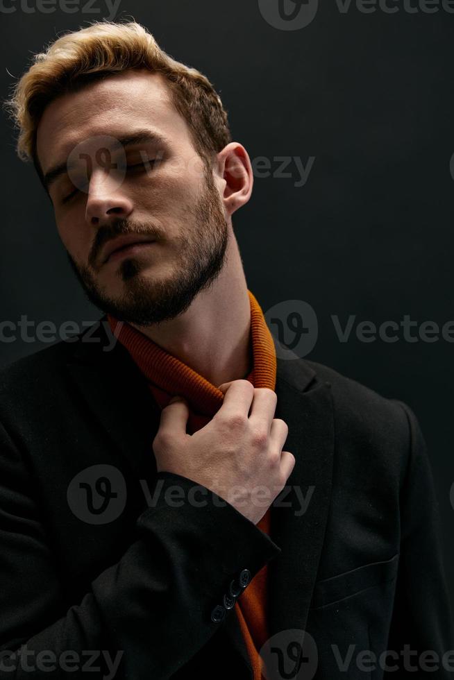 un' biondo tipo nel un arancia maglione e un' giacca detiene il suo mano vicino il suo collo su un' buio sfondo modello avvicinamento foto
