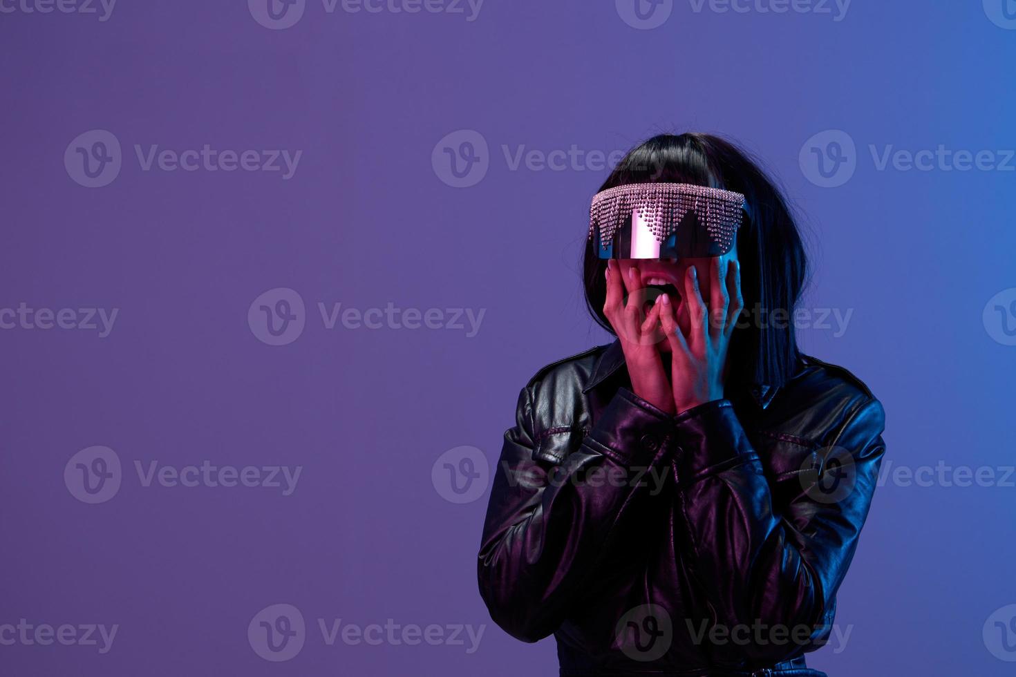 scioccato bella eccezionale brunet donna nel pelle giacca di moda speculare occhiali da sole Aperto bocca toccare guance in posa isolato nel blu viola colore leggero sfondo. neon festa cyberpunk concetto. copia spazio foto