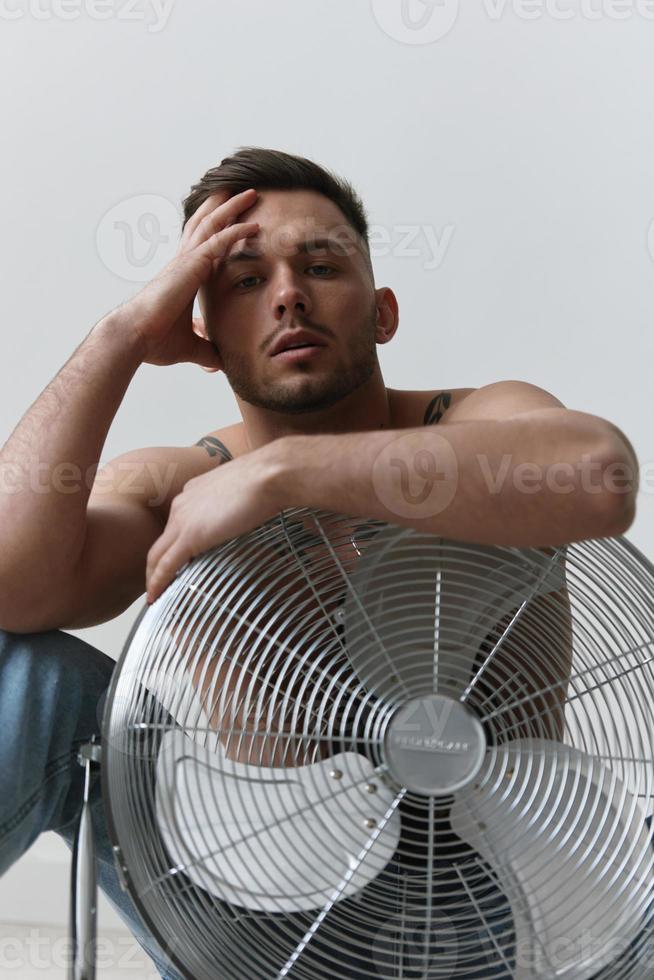 modellazione istantanee. avvicinamento ritratto di attraente nudo bello uomo nel jeans pendente su ventilatore seduta su pavimento in posa nel bianca studio sfondo. moda uomini sparare concetto. copia spazio. freddo offrire foto