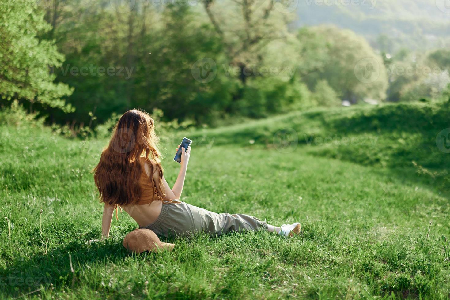 superiore Visualizza di un' donna nel un arancia superiore e verde pantaloni seduta su il estate verde erba con sua indietro per il telecamera con sua Telefono, un' giovane libero professionista studenti concetto di opera e tempo libero foto