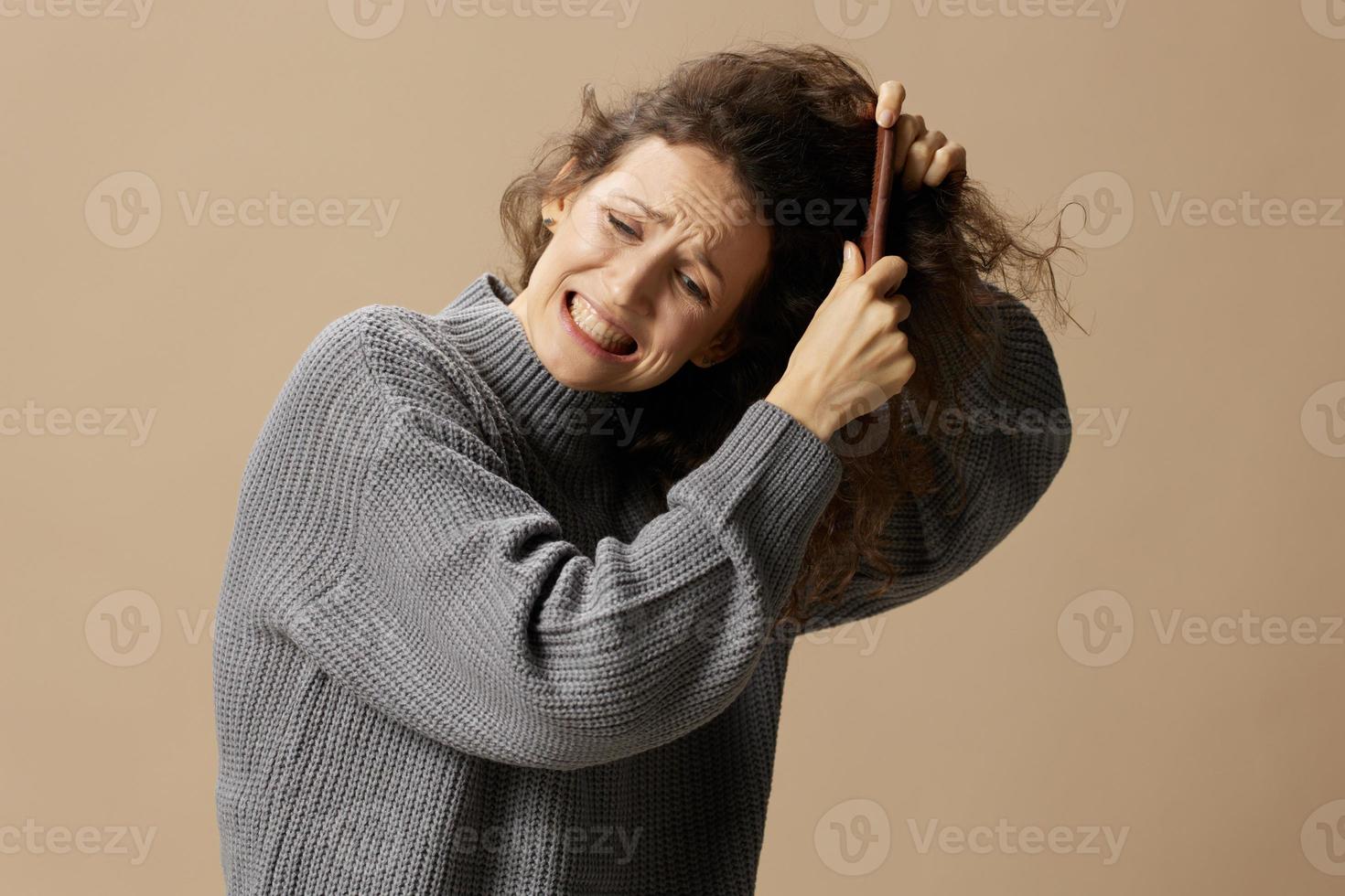 sofferenza Riccio bellissimo femmina nel grigio casuale maglione con spazzola per capelli pettine tira ingarbugliato capelli in posa isolato su al di sopra di beige pastello sfondo. problematico indisciplinato danneggiato capelli concetto. copia spazio foto