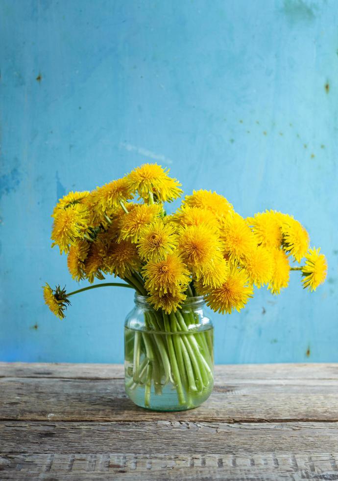 bouquet di denti di leone in vaso di vetro sul tavolo di legno con sfondo azzurro foto