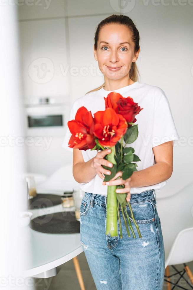 ritratto di giovane donna nel bianca maglietta con mazzo di rosso fiori nel mani vicino finestra nel luminosa cucina a casa foto