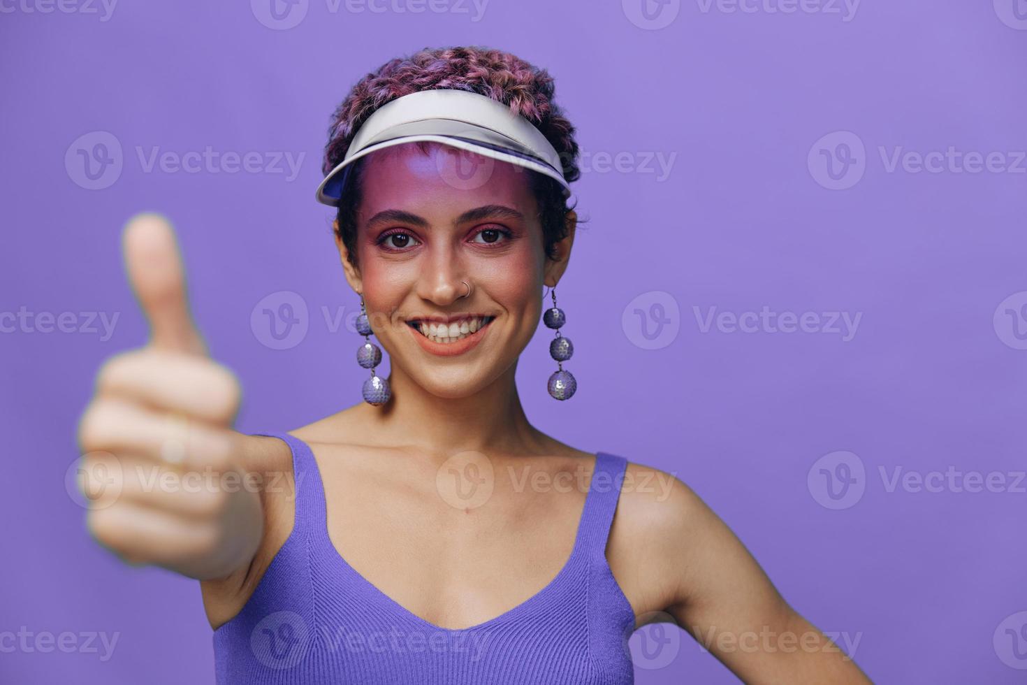 ritratto di un' sportivo moda donna in posa sorridente con denti e puntamento un' dito a il telecamera nel un' viola yoga tuta da ginnastica e un' trasparente berretto su un' viola monocromatico sfondo foto