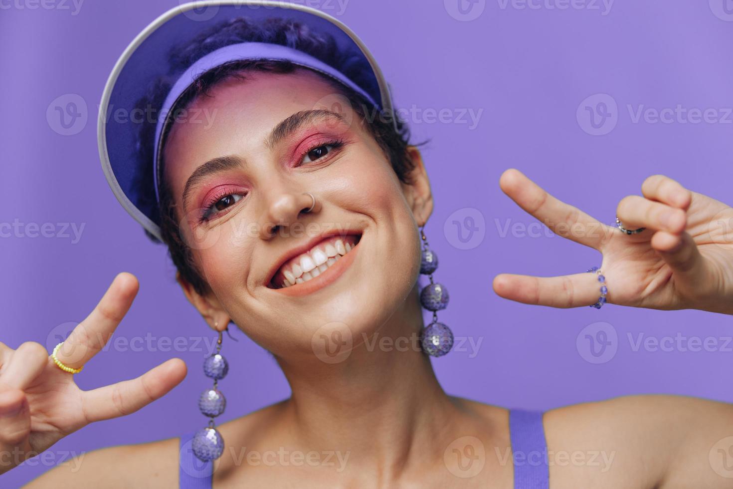ritratto di un' sportivo moda donna in posa sorridente con denti a il telecamera con elegante orecchini nel sua orecchie nel un' viola yoga tuta da ginnastica e un' trasparente berretto su un' viola monocromatico sfondo foto