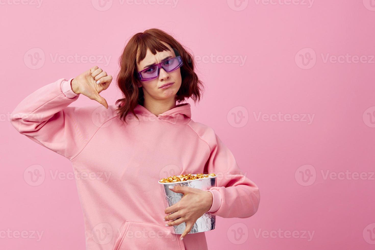 freddo film. confuso carino testa Rossa signora nel rosa felpa con cappuccio occhiali da sole con Popcorn in posa isolato su al di sopra di rosa studio sfondo mostrare copia spazio striscione. moda cinema concetto. divertimento offrire foto