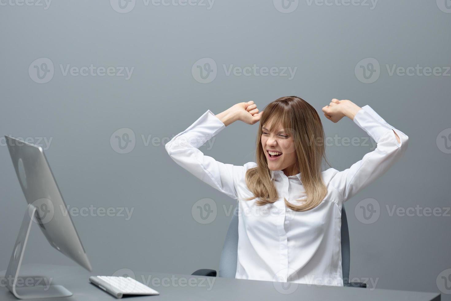 felicissimo bella bionda donna d'affari lavoratore libero professionista utilizzando del desktop computer aumentare mani su festeggiare vincere nel grigio moderno ufficio. a distanza lavoro, tecnologia e carriera professione concetto. copia spazio foto