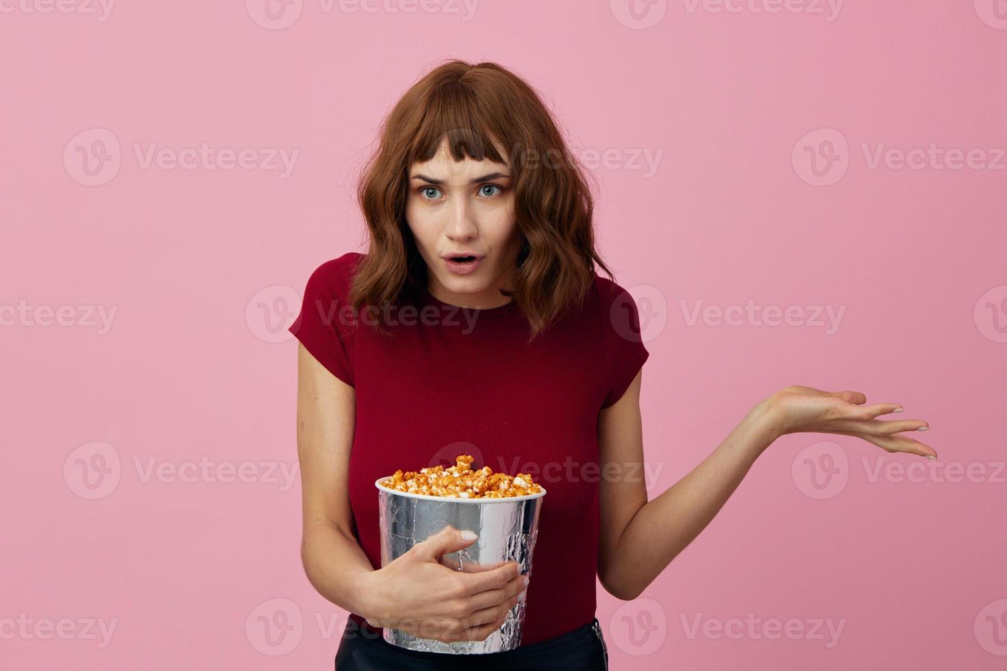 eccitato scioccato carino testa Rossa signora nel rosso maglietta con Popcorn pronto per film sera in posa isolato su al di sopra di rosa studio sfondo. copia spazio striscione. moda cinema concetto. divertimento offrire foto