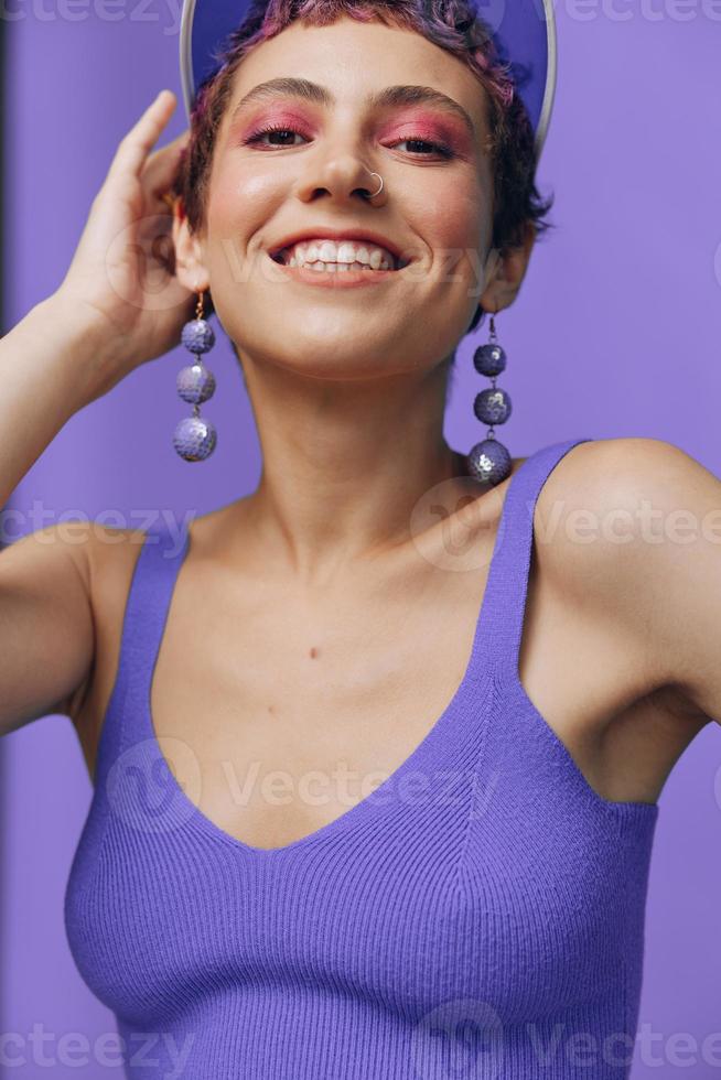 ritratto di un' sportivo moda donna in posa sorridente con denti a il telecamera con elegante orecchini nel sua orecchie nel un' viola yoga tuta da ginnastica e un' trasparente berretto su un' viola monocromatico sfondo foto