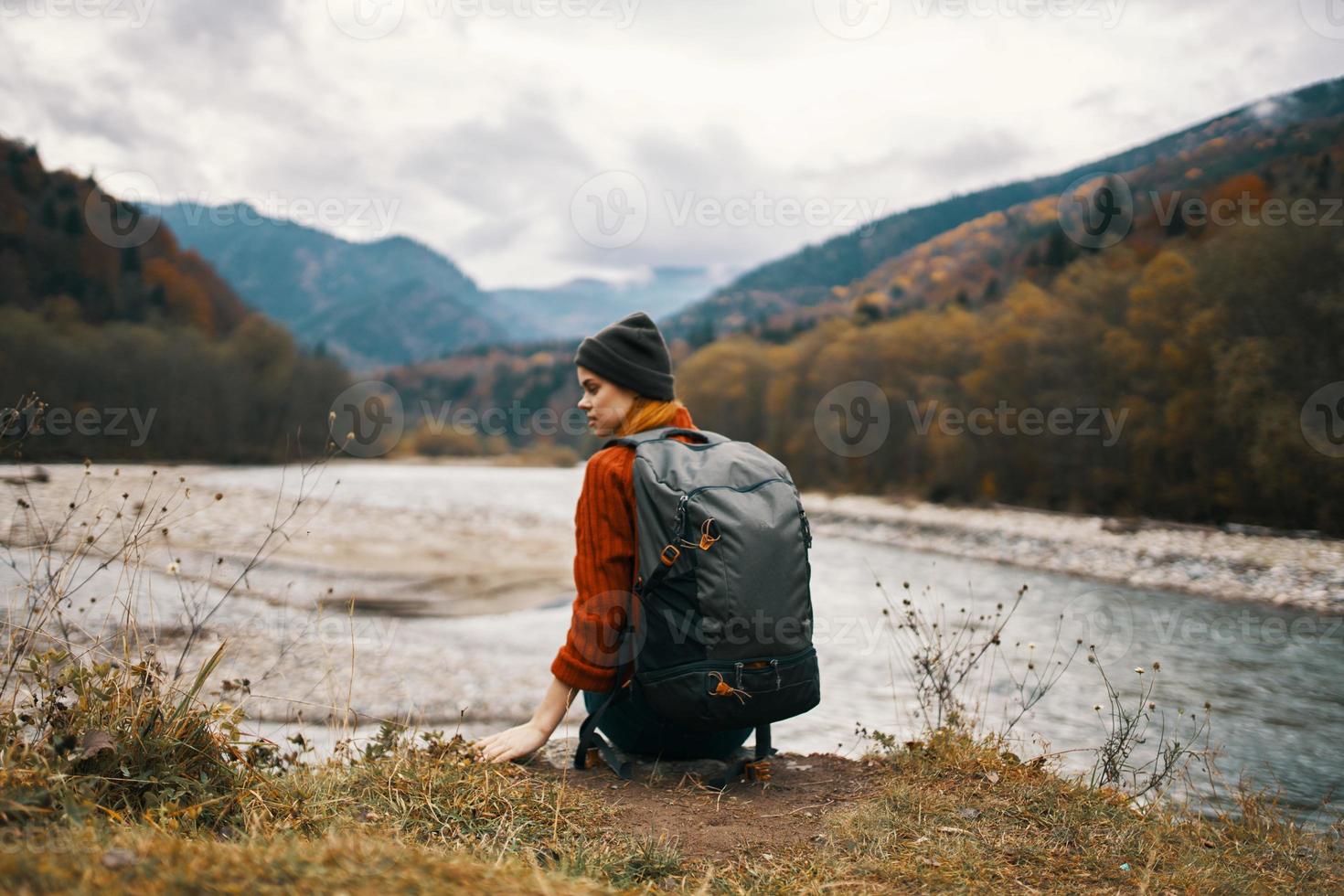 donna con un' zaino e nel un' giacca con un' cappello su il fiume banca nel il montagne nel natura foto