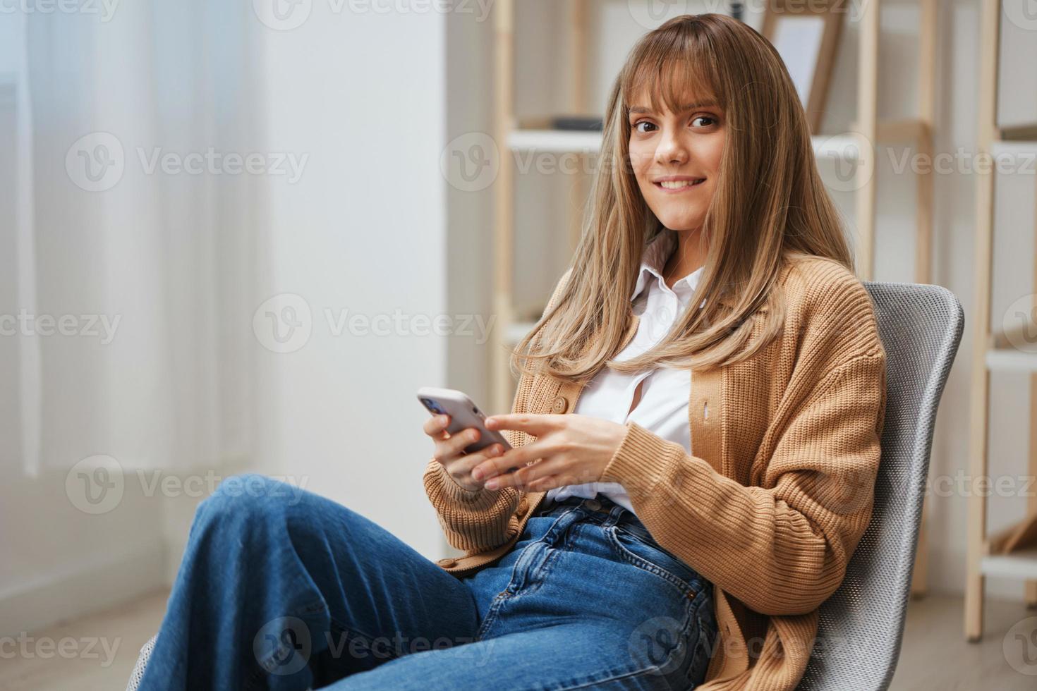 contento carino giovane bionda signora nel caldo maglione fare in linea shopping uso Telefono sembra a telecamera seduta nel poltrona a casa. pausa a partire dal opera, prendere un' rompere, sociale media nel gratuito tempo concetto. copia spazio foto