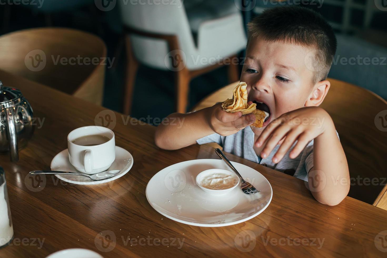 poco e carino ragazzo avendo prima colazione nel bar. mangiare Pancakes con acida crema foto
