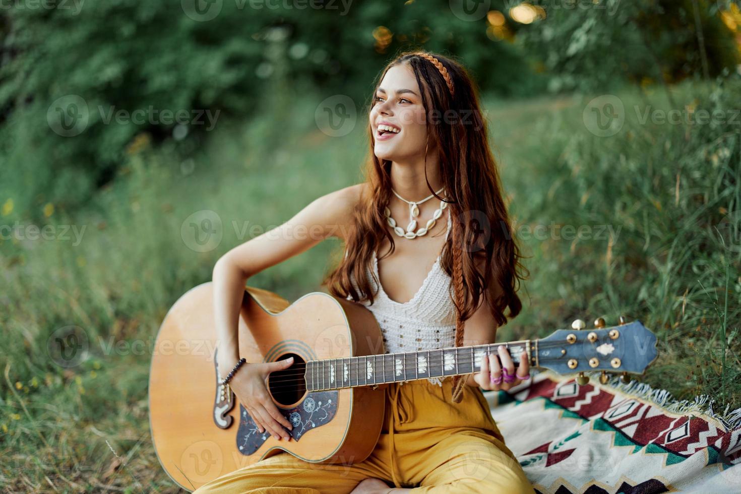 giovane femmina hippie artista giochi chitarra e canta canzoni nel eco-friendly capi di abbigliamento seduta su il terra al di fuori nel natura nel il autunno guardare su a il tramonto foto