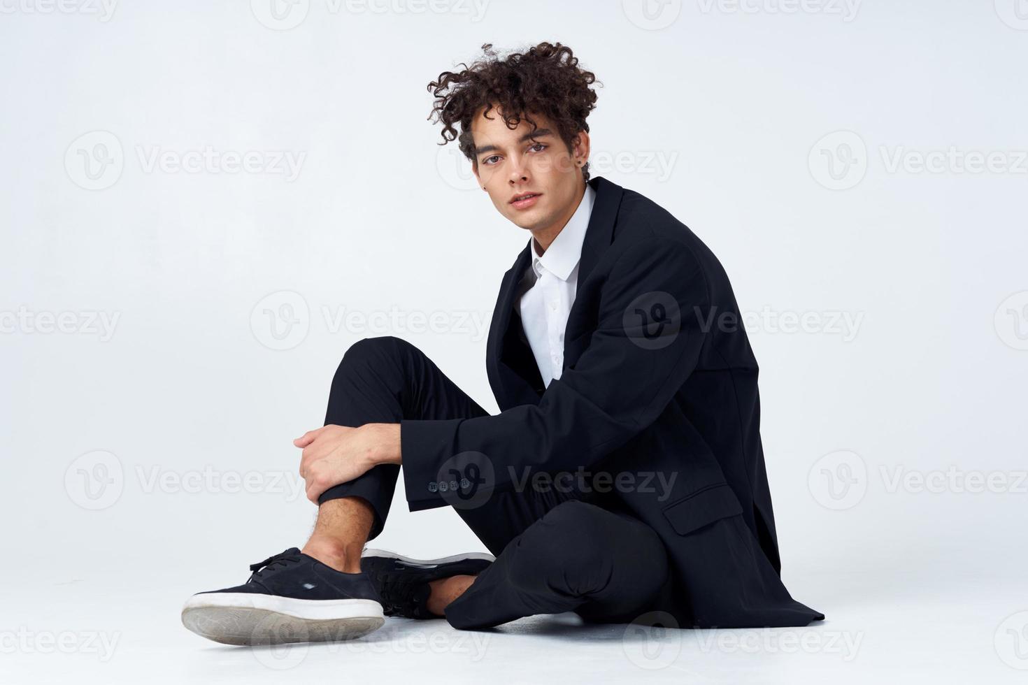 elegante giovane uomo nel completo da uomo si siede su il pavimento e scarpe da ginnastica Riccio capelli moda foto