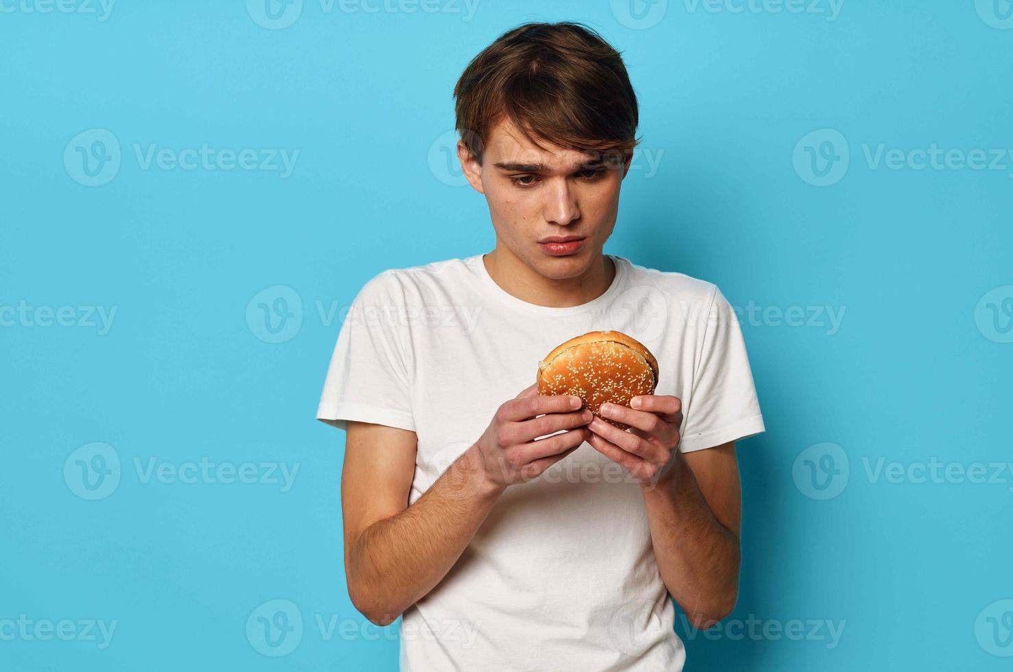 un' uomo nel un' bianca maglietta con un' Hamburger nel il suo mani pranzo blu sfondo foto