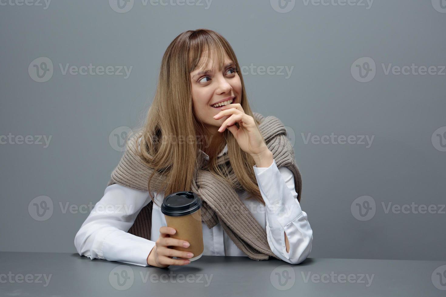 sorridente sognare giovane bionda alunno signora libero professionista nel caldo maglione con porta via caffè reclinare su mano sembra su nel grigio moderno casa ufficio. caffè rompere amante concetto. copia spazio. freddo offrire foto