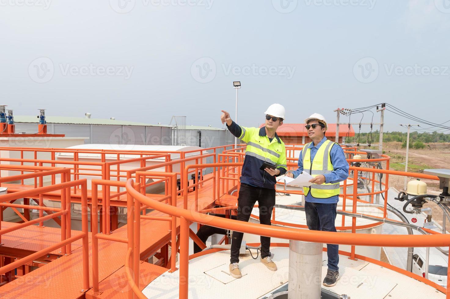 Due pesante industriale ingegneri in piedi nel un' standard acqua trattamento pianta uso digitale tavoletta computer e chattare. asiatico industria professionisti foto