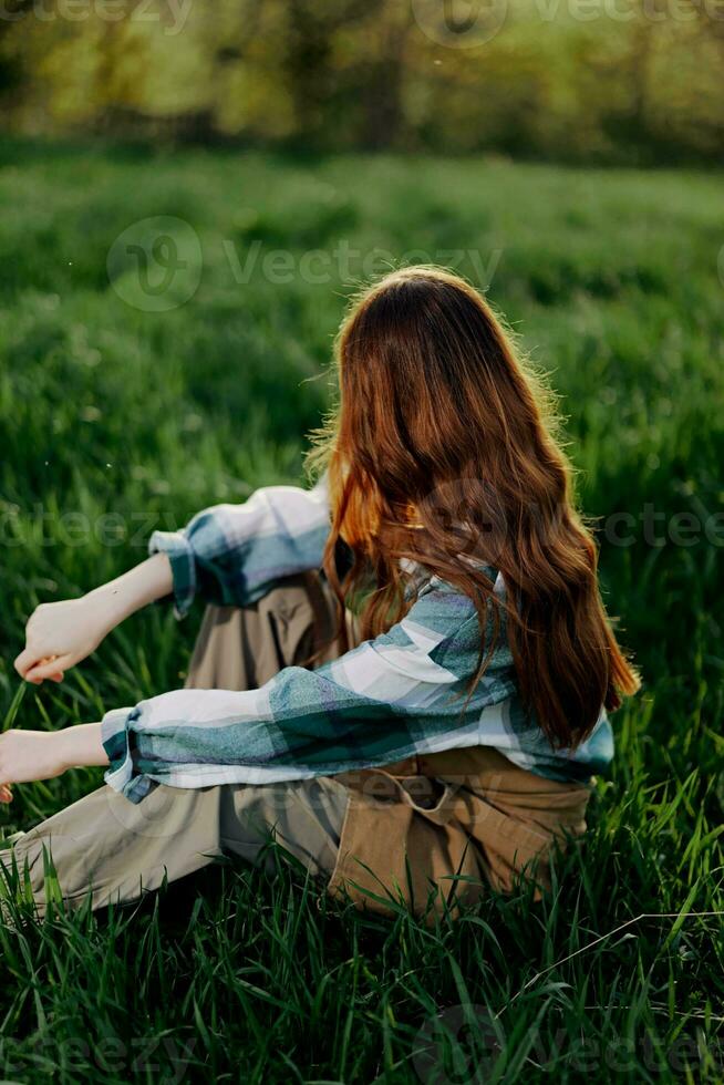 un' giovane, bellissimo donna salotti su il verde erba nel il parco indossare sneaker pantaloni e un' plaid camicia e sembra su in il ambientazione sole foto