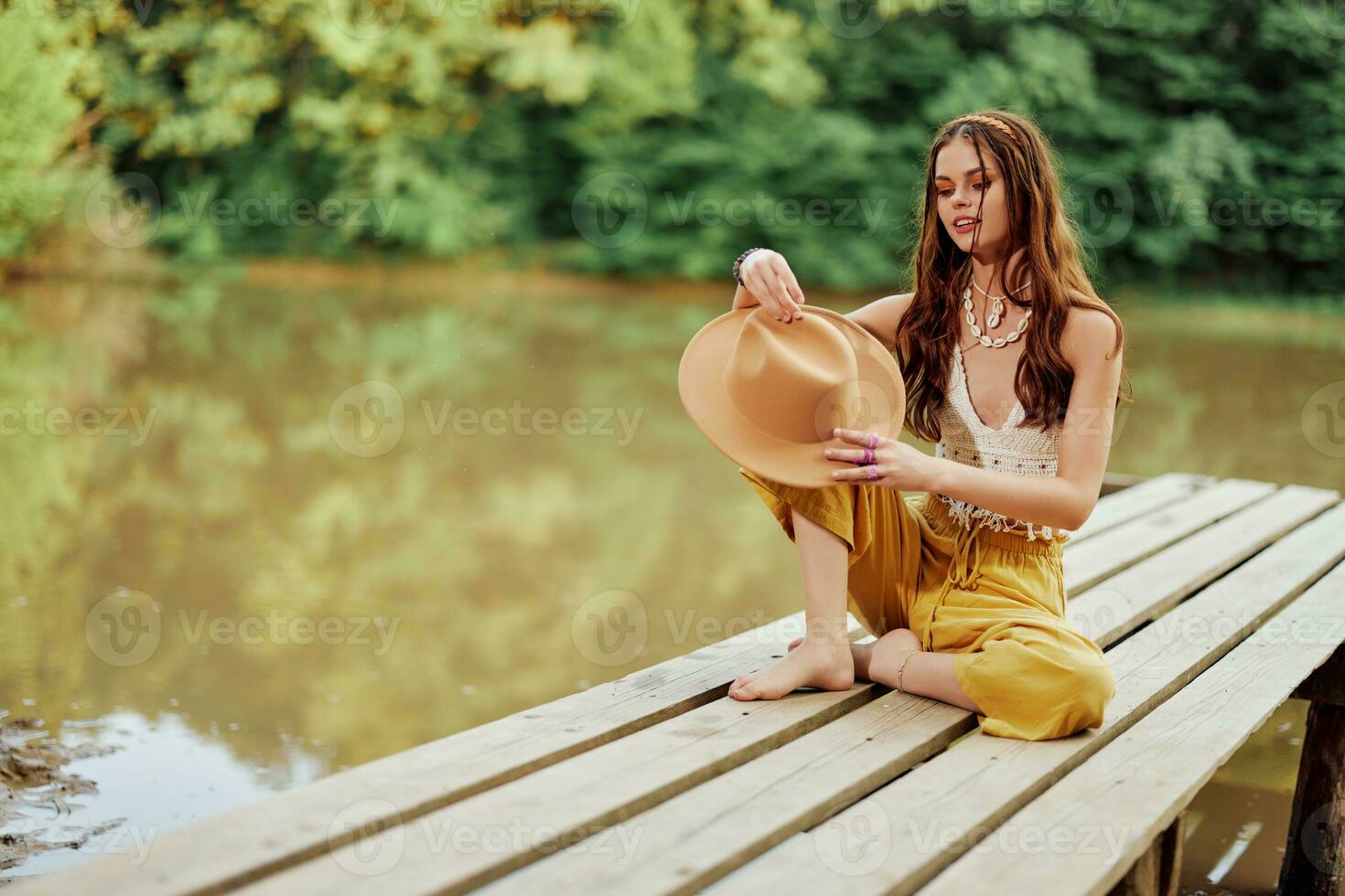 un' giovane hippie donna si siede su un' lago ponte indossare elegante eco Abiti e sorridente foto