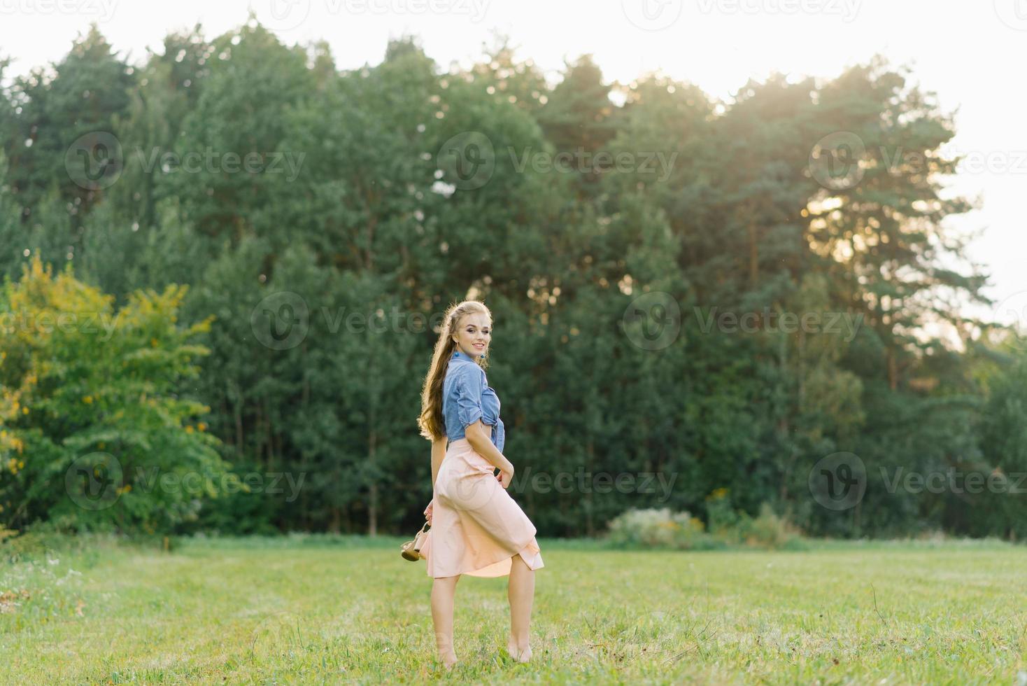 giovane caucasico donna o ragazza nel un' estate vestito e un' denim camicia è a piedi su il erba, Tenere scarpe nel sua mani, rilassante al di fuori il città su un' fine settimana nel estate foto