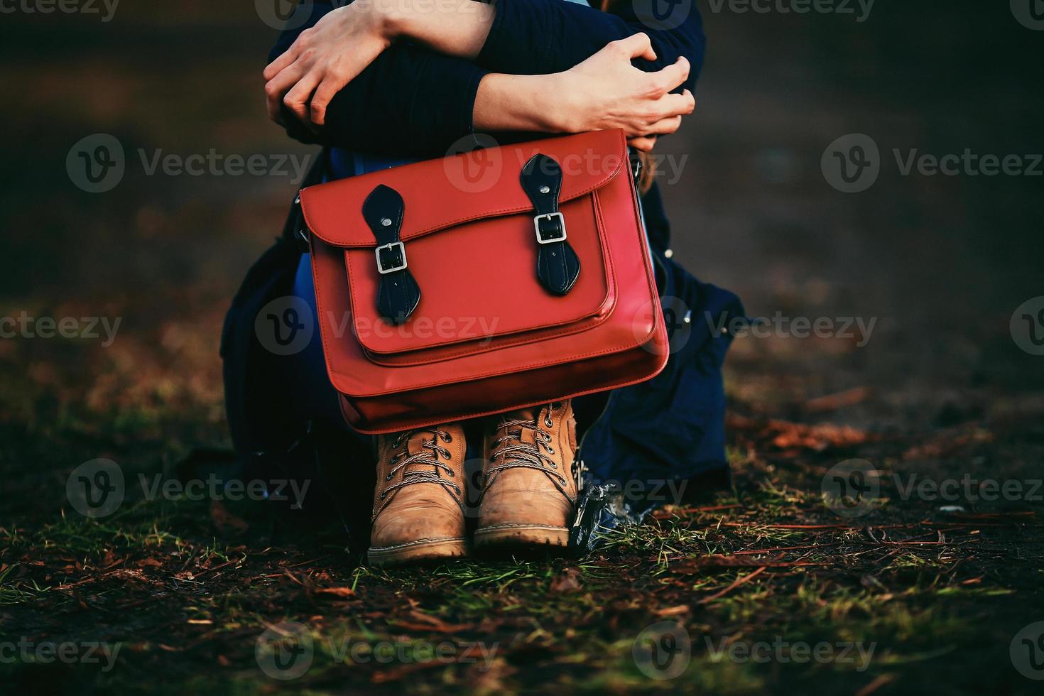 ragazza alla moda in scarpe marroni e un cappotto caldo seduto nel parco con una borsa rossa. foto