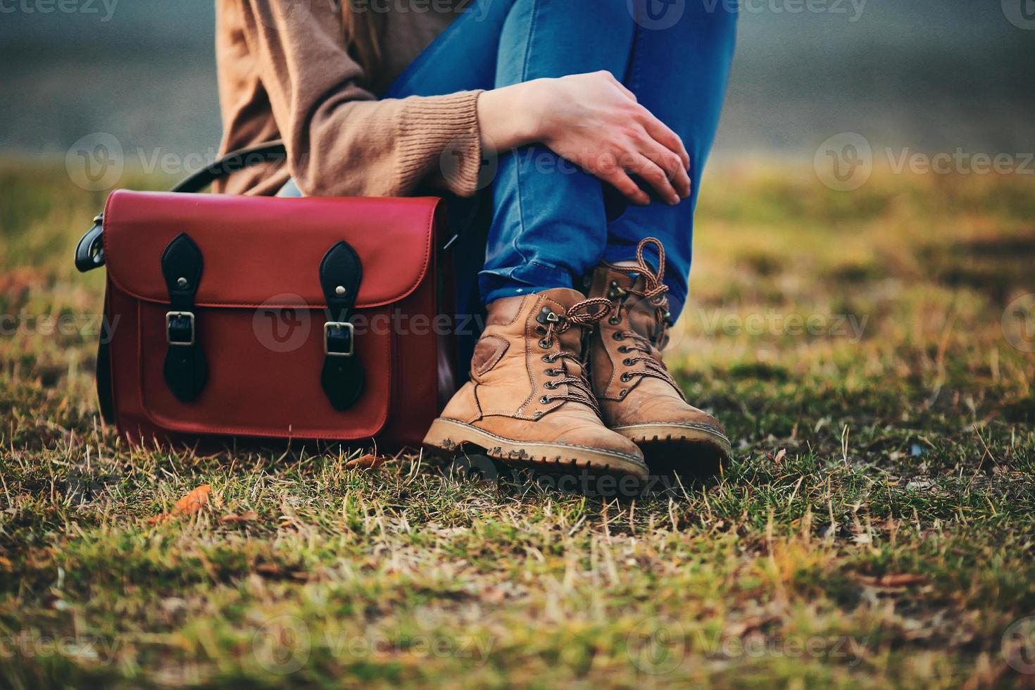 ragazza alla moda in scarpe marroni e un cappotto caldo seduto nel parco con una borsa rossa. foto