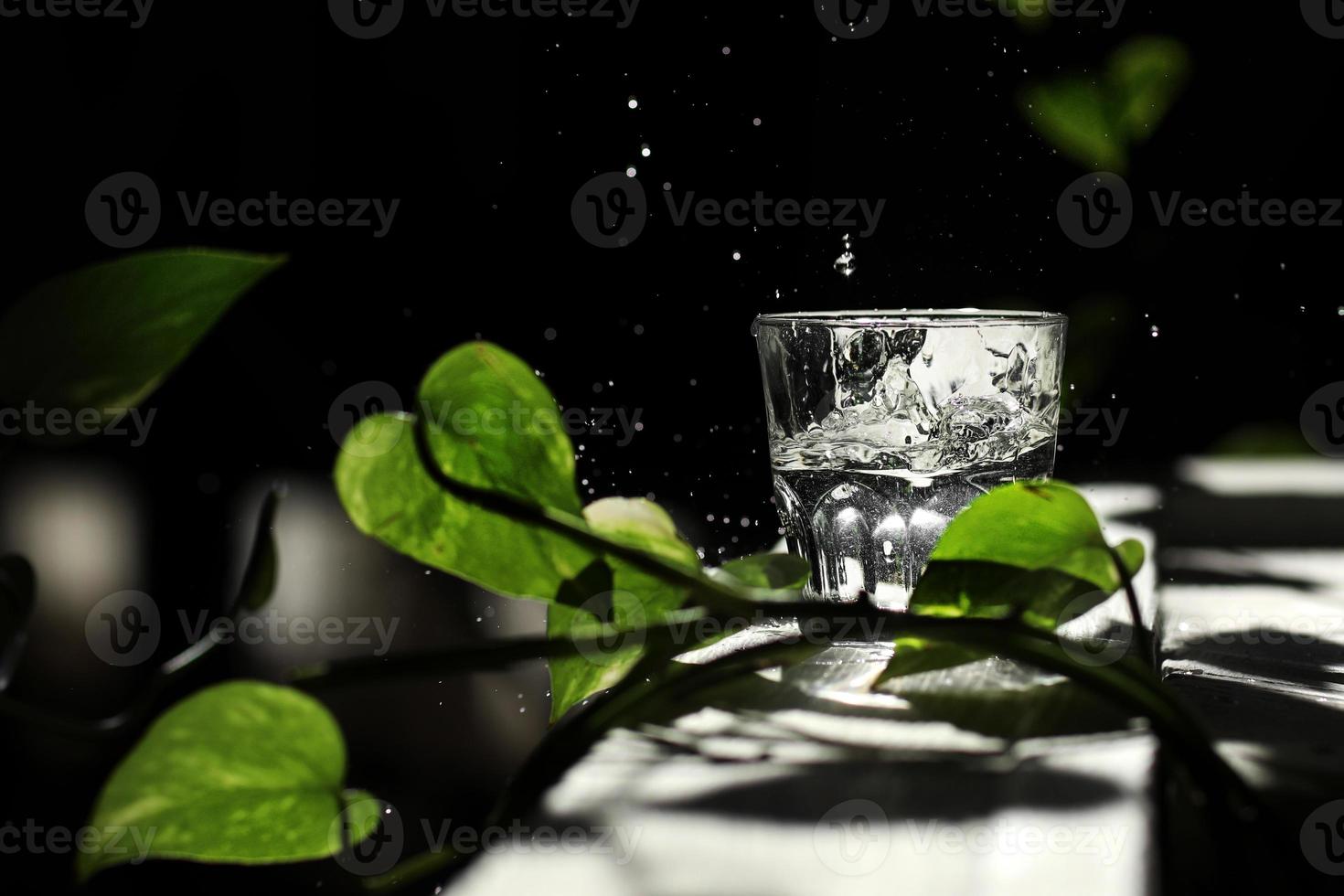 un bicchiere d'acqua su uno sfondo scuro tra le foglie verdi. concetto di eco. acqua versata da un bicchiere. gocce d'acqua su uno sfondo nero foto