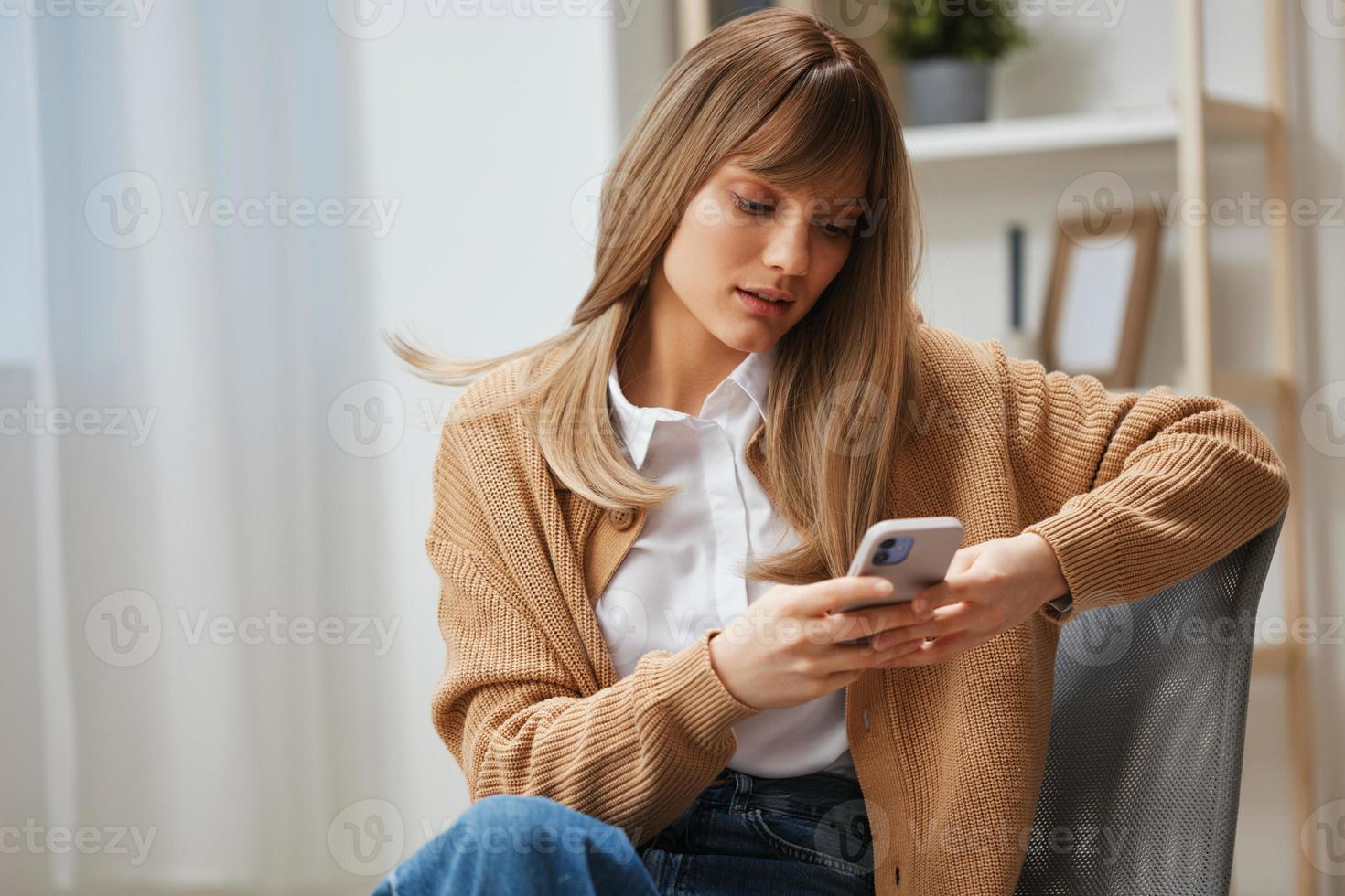 focalizzata grave giovane bionda signora nel caldo maglione digitando Messaggio uso Telefono seduta nel poltrona a moderno casa interno. pausa a partire dal opera, prendere un' rompere, sociale media nel gratuito tempo concetto. copia spazio foto