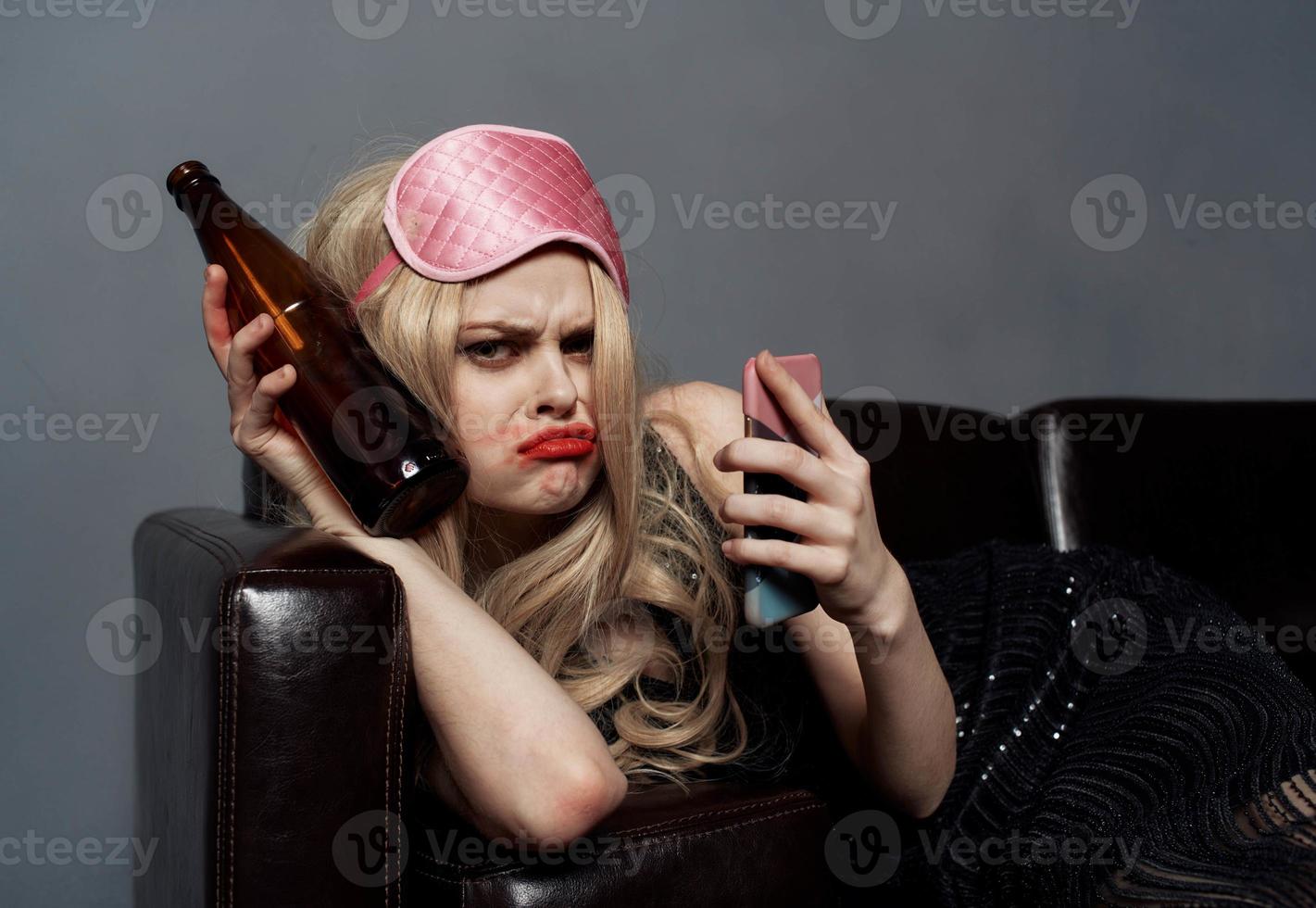 un' donna nel un' rosa maschera bugie su il divano con un' bottiglia di birra e un' Telefono nel sua mano foto