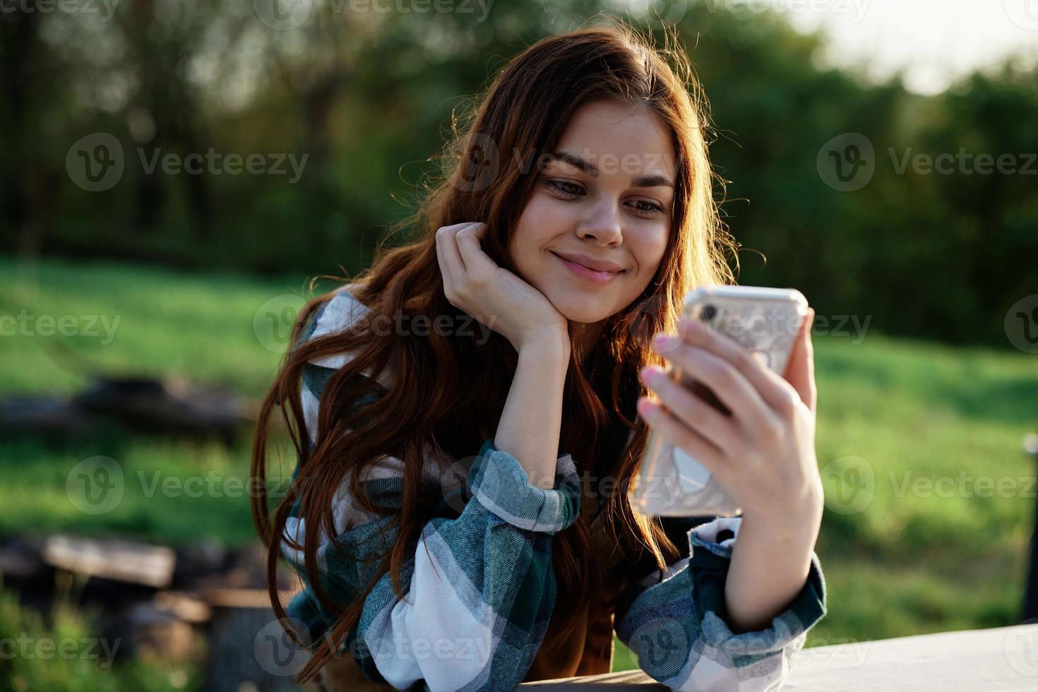 un' bellissimo donna rilassante e Lavorando su sua Telefono seduta nel natura nel il parco tra il alberi sorridente e Tenere sua smartphone nel sua mano illuminato di il luminosa tramonto leggero foto