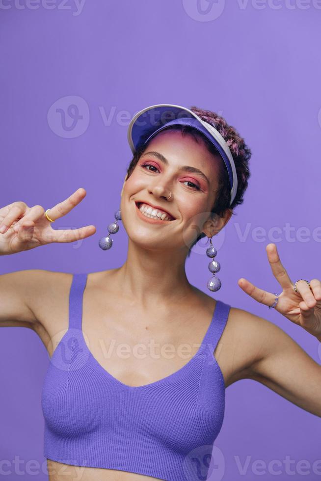 ritratto di un' sportivo moda donna in posa sorridente con denti a il telecamera con elegante orecchini nel sua orecchie nel un' viola yoga tuta da ginnastica e un' trasparente berretto su un' viola monocromatico sfondo foto