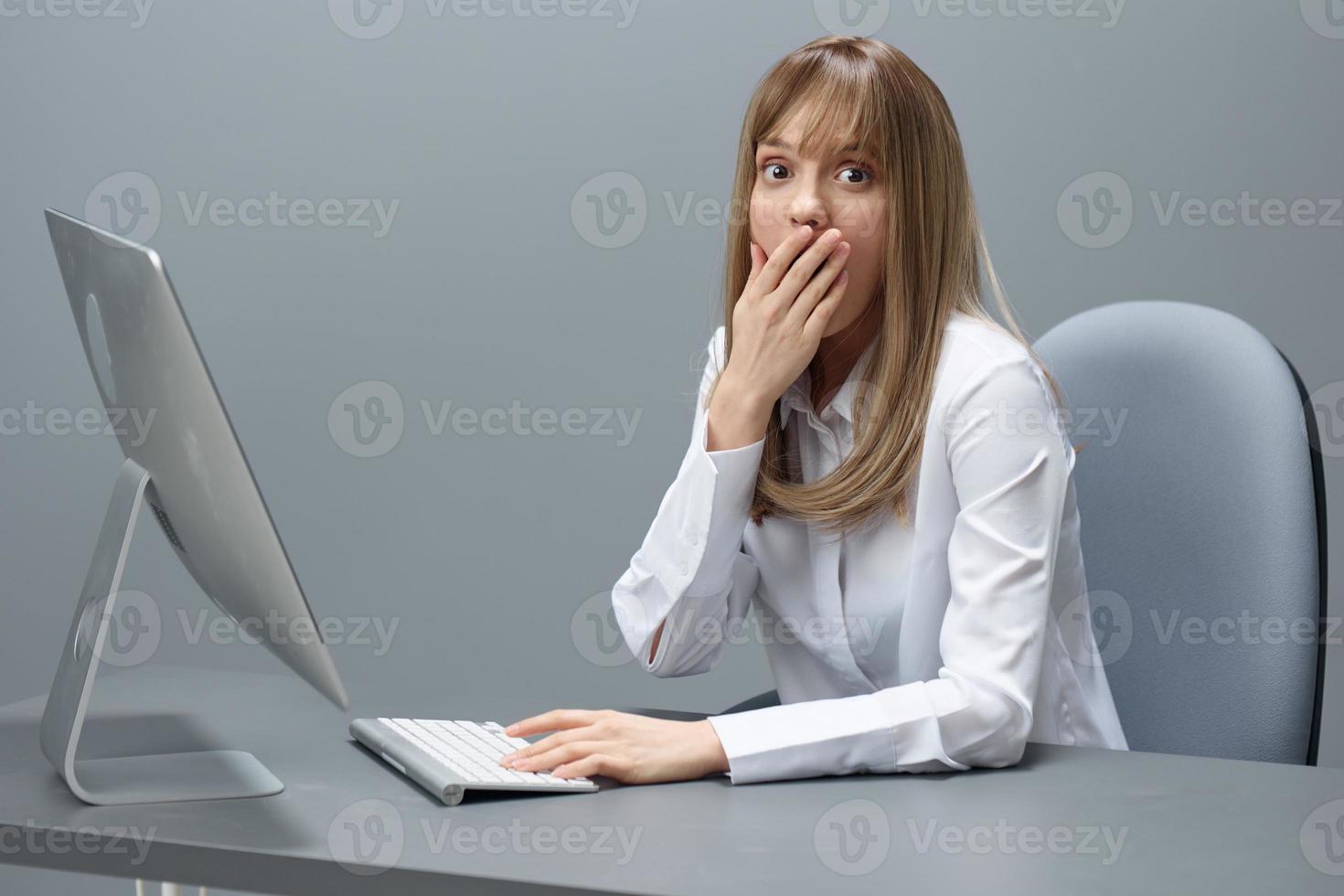 scioccato bella bionda donna d'affari lavoratore libero professionista utilizzando del desktop computer vicino bocca di mano seduta a posto di lavoro nel grigio moderno ufficio. a distanza lavoro e carriera professione concetto. copia spazio foto