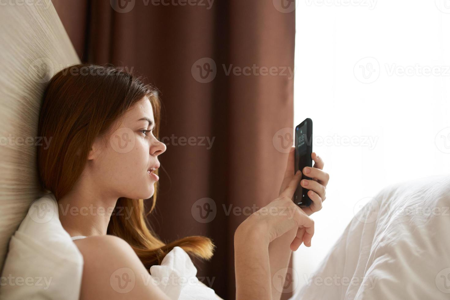 donna con Telefono nel mano nel letto vicino il finestra, tenda foto