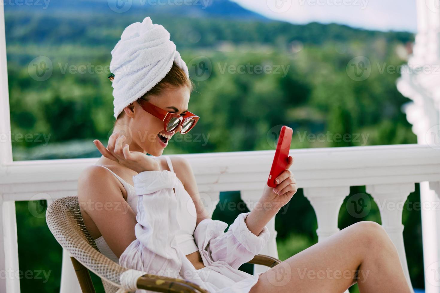 attraente giovane donna nel occhiali da sole si siede su il balcone e usi il Telefono pigro mattina foto