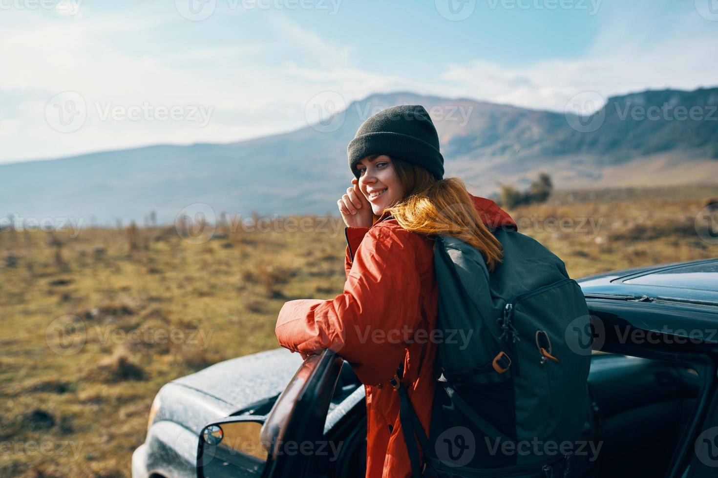 donna nel un' giacca con un' zaino e un' berretto sta vicino il auto su natura nel il montagne nel estate foto