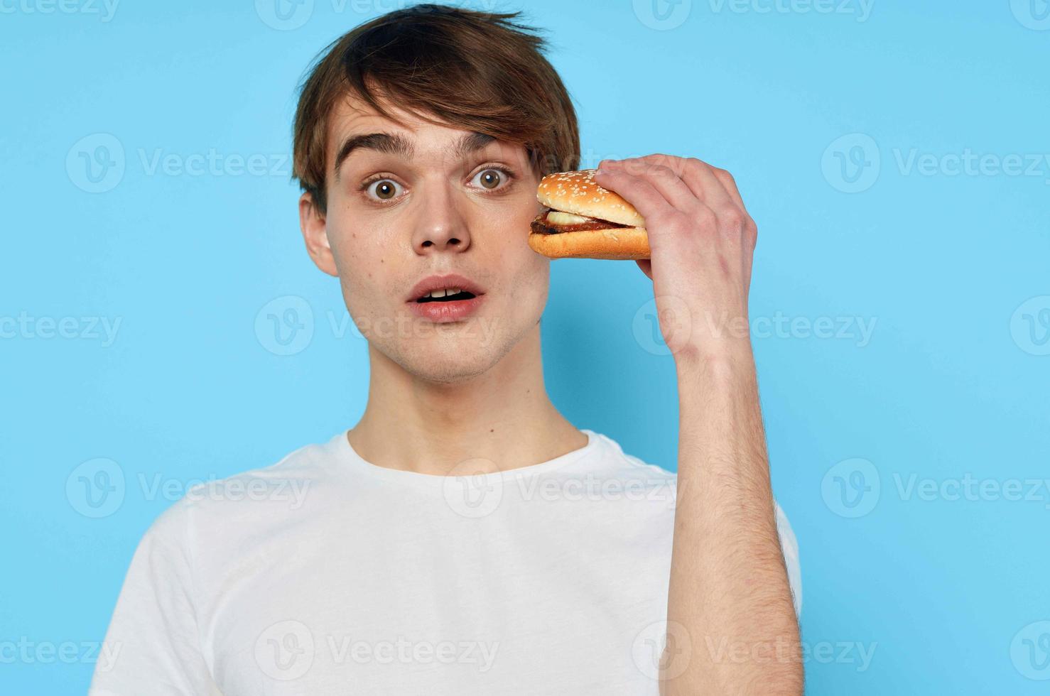 un' uomo nel un' bianca maglietta con un' Hamburger nel il suo mani pranzo blu sfondo foto