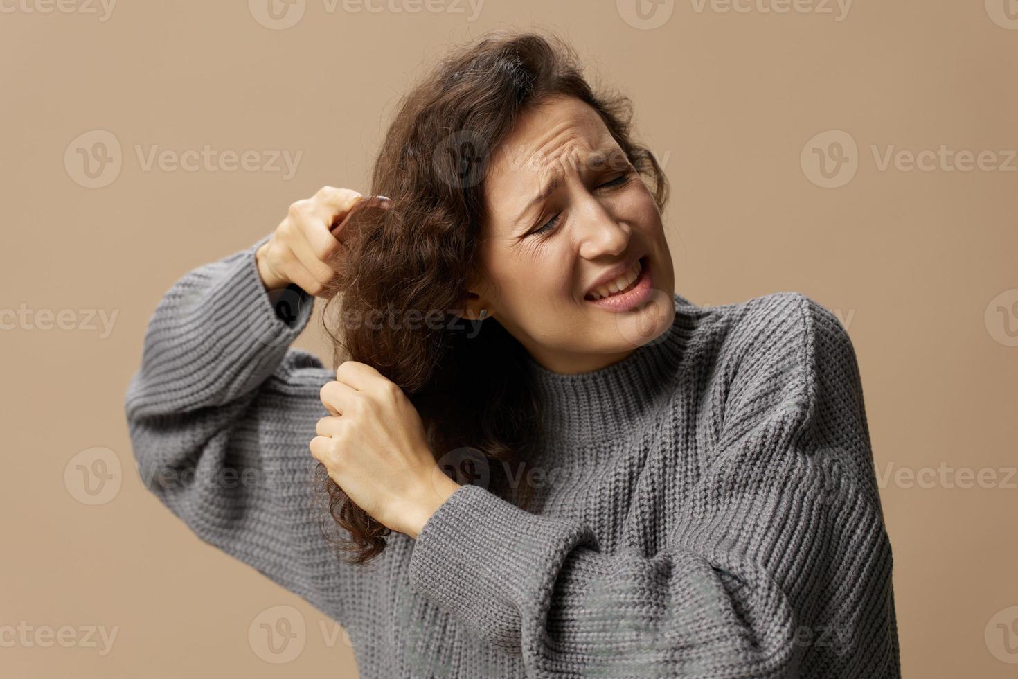 danneggiato capelli concetto. triste sofferenza irritata Riccio bellissimo donna nel grigio casuale maglione con spazzola per capelli pettine tira ingarbugliato capelli in posa isolato su al di sopra di beige pastello sfondo. copia spazio foto