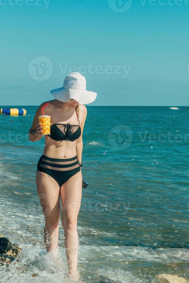 un' bellissimo ragazza nel un' costume da bagno e un' cannuccia cappello passeggiate lungo il riva del mare su un' soleggiato estate giorno, nel sua mani è un' bicchiere con un' cannuccia e un' freddo bevanda e un' telefono foto