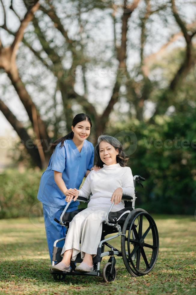 anziano asiatico anziano donna su sedia a rotelle con asiatico attento badante e incoraggiare paziente, a piedi nel giardino. con cura a partire dal un' badante e anziano Salute assicurazione. foto