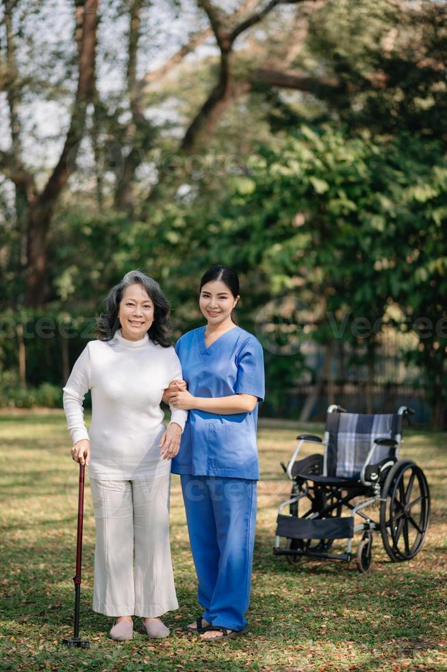 asiatico attento badante o infermiera e il contento paziente nel un' sedia a rotelle siamo a piedi nel il giardino per Aiuto e incoraggiare e riposo il tuo mente con verde natura. Aiuto supporto te stesso per imparare per camminare. foto