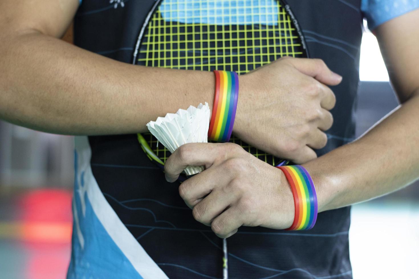 badminton giocatore indossa arcobaleno braccialetti e Tenere racchetta e bianca volano nel davanti di il netto prima servendo esso per giocatore nel un altro lato di il Tribunale, concetto per lgbt persone attività. foto